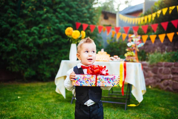 Сюрприз на день рождения. Мальчик держит коробку с подарком во дворе на фоне праздничного стола с тортом. одетый в черный костюм — стоковое фото