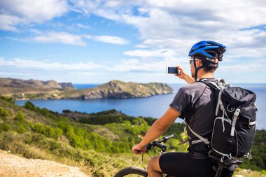 genç bir adam üzerinde bir dağ bisikleti rotaları İspanya've Akdeniz Costa Brava kayalık kıyılarının arka planda beyaz bir cep telefonu üzerinde bir fotoğrafı çeker. Mavi bir kask kafası ve bir b
