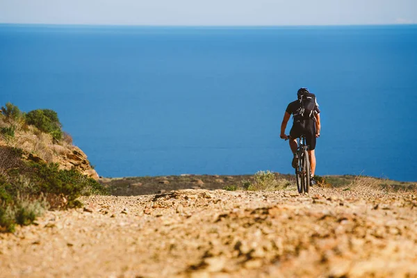 Un joven montando una bicicleta de montaña en una ruta en bicicleta en España en la carretera contra el fondo del mar Mediterráneo. Vestido con un casco, uno oscuro y una mochila negra — Foto de Stock
