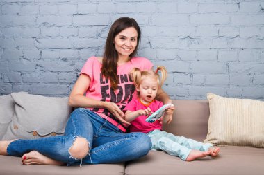 parlak renkler, renkli giysi odasında Dressed kanepede oturan iki yaşındaki kızı ile oynamak için bir akıllı telefon güzel genç anne kullanır