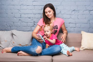 parlak renkler, renkli giysi odasında Dressed kanepede oturan iki yaşındaki kızı ile oynamak için bir akıllı telefon güzel genç anne kullanır