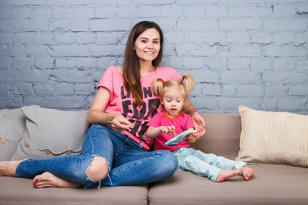 Bela jovem mãe usa um smartphone para brincar com uma filha de dois anos sentada no sofá no quarto Vestida com cores brilhantes, roupas coloridas — Fotografia de Stock