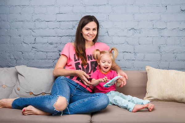 parlak renkler, renkli giysi odasında Dressed kanepede oturan iki yaşındaki kızı ile oynamak için bir akıllı telefon güzel genç anne kullanır