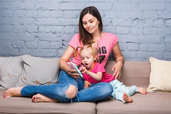 parlak renkler, renkli giysi odasında Dressed kanepede oturan iki yaşındaki kızı ile oynamak için bir akıllı telefon güzel genç anne kullanır