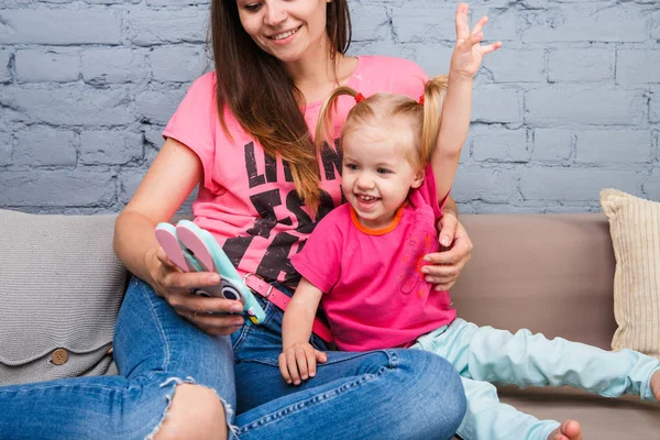 Belle jeune mère utilise un smartphone pour jouer avec une fille de deux ans assise sur le canapé dans la chambre Habillée de couleurs vives, vêtements colorés — Photo