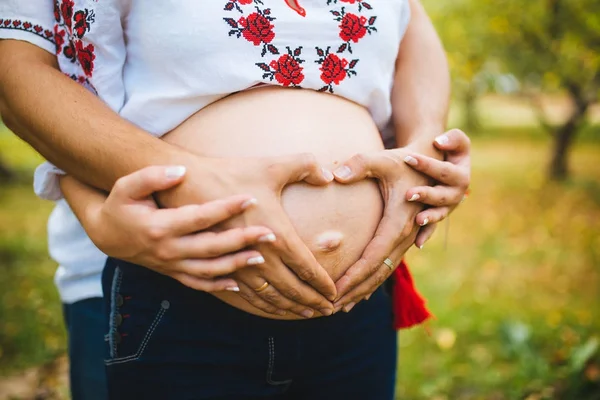 Έγκυος κοιλιά με δάχτυλα καρδιά σύμβολο. Όμορφη νεαρή έγκυος γυναίκα και συζύγου μαζί χαϊδεύει την κοιλιά της εγκύου. Εγκυμοσύνη. Έννοια της αγάπης. Μαμά και ο μπαμπάς σε αναμονή για το νεογέννητο μωρό. — Φωτογραφία Αρχείου