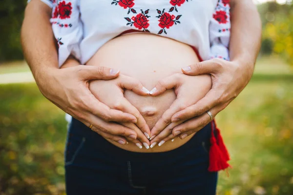 Έγκυος κοιλιά με δάχτυλα καρδιά σύμβολο. Όμορφη νεαρή έγκυος γυναίκα και συζύγου μαζί χαϊδεύει την κοιλιά της εγκύου. Εγκυμοσύνη. Έννοια της αγάπης. Μαμά και ο μπαμπάς σε αναμονή για το νεογέννητο μωρό. — Φωτογραφία Αρχείου
