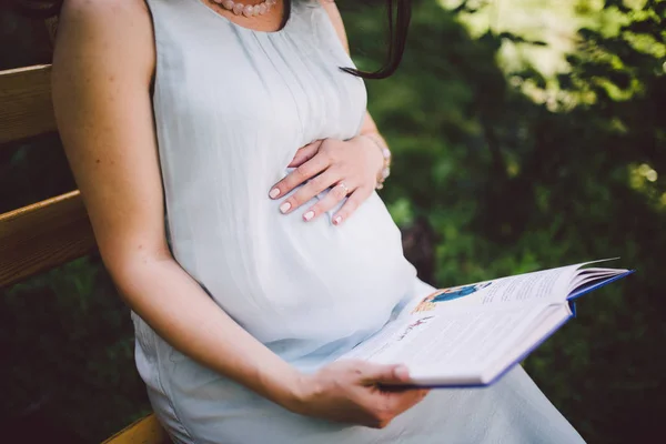 Wanita hamil membaca sebuah buku di bangku dan memegang tangannya di perutnya pada musim panas — Stok Foto