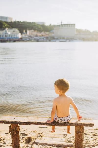 Lindo bebé niño en un banco al aire libre en la playa con vistas al mar o al agua del mar en un día soleado . —  Fotos de Stock