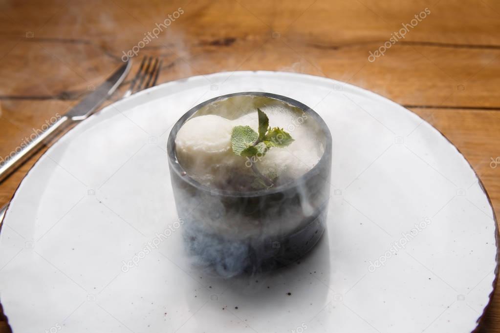 ice cream sweet dessert on a white plate on a wooden table in an Italian retor. Ingredients white ice cream with mint and cocoa in a transparent tequal glass with smoke.