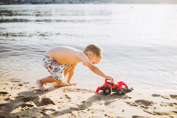 Мальчик играет на пляже рядом с речной игрушкой красный трактор — стоковое фото