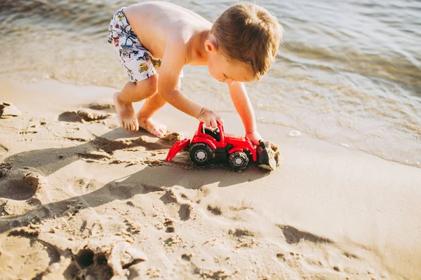 Niño niño jugando en la playa cerca del río juguete rojo tractor —  Fotos de Stock