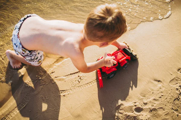 Мальчик играет на пляже рядом с речной игрушкой красный трактор — стоковое фото