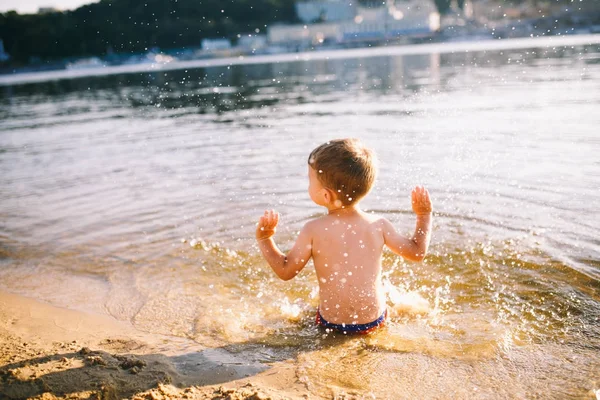 Niño niño jugando hace salpicaduras, bate las manos en el agua en el río al atardecer —  Fotos de Stock