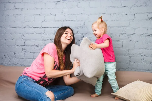 Máma a dcera hrát na gauči s polštáře boj. Oblečený ve světlé stylové oblečení v místnosti — Stock fotografie