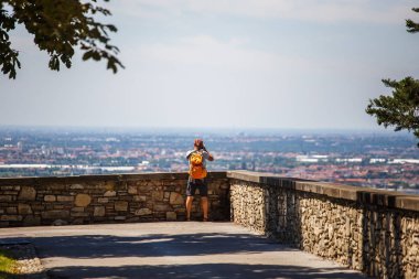 turist adam ile görüntüleme platformu Bergamo İtalya sırtında duruyor ve şort, sandalet ve turuncu bir sırt çantası fotoğraf makinesi üzerinde bir fotoğraf çeker.