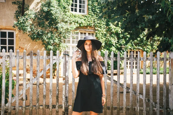 Belle fille brune sexy aux yeux marron dans une robe noire et un chapeau noir avec de gros bords sur fond de clôture en bois et une vieille maison dans un village en Bourgogne en france — Photo