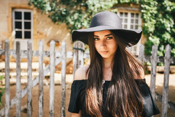 Hermosa chica sexy morena con ojos marrones en un vestido negro y un sombrero negro con grandes alas contra el telón de fondo de una valla de madera y una casa vieja en un pueblo en Borgoña en Francia — Foto de Stock