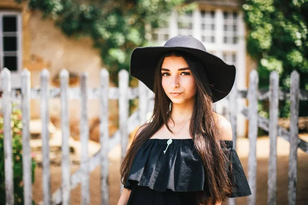 Hermosa chica sexy morena con ojos marrones en un vestido negro y un sombrero negro con grandes alas contra el telón de fondo de una valla de madera y una casa vieja en un pueblo en Borgoña en Francia — Foto de Stock