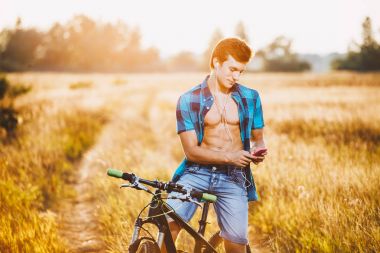 Hoş bir genç adam elinde bir akıllı telefon kullanan ve gün batımında alanında bir bisiklet üzerinde otururken kulaklık müzik dinler. Şık elbise gömlek ve kot pantolon giymiş.