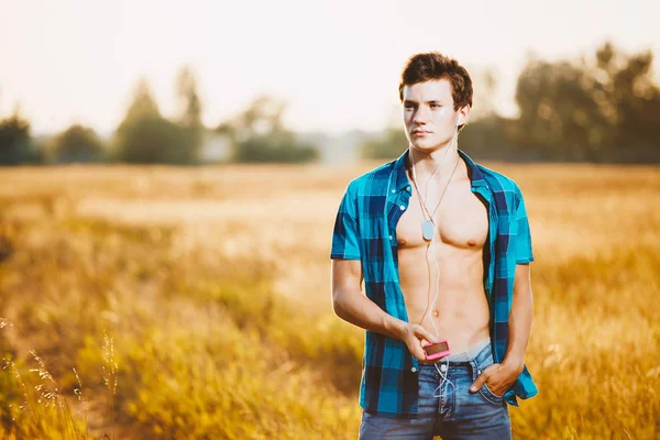 Un guapo joven caucásico con un cuerpo musculoso vestido con una camisa azul desabotonada y pantalones cortos de mezclilla escuchando música en los auriculares, sosteniendo un smarfon en su mano. En el campo amarillo al atardecer o — Foto de Stock