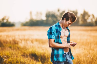 beyaz kulaklık müzik dinlemek ve bir akıllı telefon elinde tutan kas gövde ile yakışıklı bir genç adam. O parlak mavi tişört ve kot şort giymiş, şehir dışında bir alanda