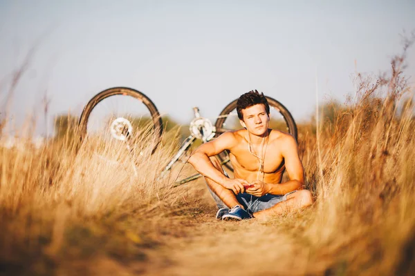 Um cara musculoso bonito com um tronco nu sentado perto de uma bicicleta no campo em um campo descansando, ouvindo música e segurando um gadget na mão, smarfon . — Fotografia de Stock