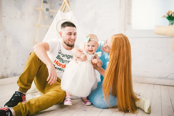 Een gelukkig gezin van drie. Vader, moeder, kind één jaar oud meisje in jurk spelen, lachen, glimlachen in lichte kamer. Zonnig weer — Stockfoto