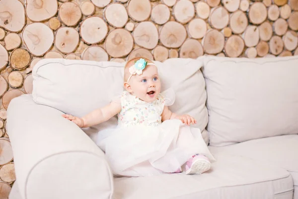 Pequena menina de olhos azuis loira em um vestido de tule branco com uma decoração em sua cabeça jogando e se alegrando em um sofá bege em uma sala com parede de madeira decorativa na forma de cânhamo — Fotografia de Stock