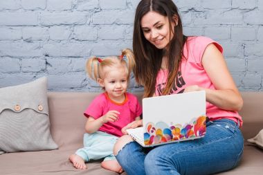 Genç anne ve iki yaşında sarışın kızı ile parlak baskı kapalı kanepede oturan dizüstü bilgisayar laptop beyaz kullanın