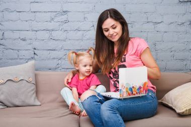 Genç anne ve iki yaşında sarışın kızı ile parlak baskı kapalı kanepede oturan dizüstü bilgisayar laptop beyaz kullanın