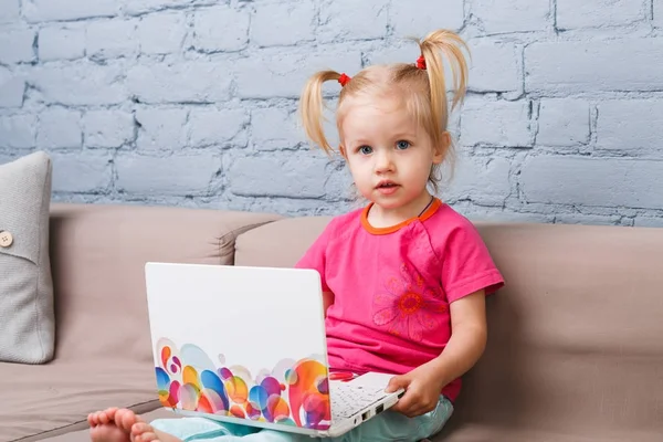 Kız çocuk üç yaşında kapalı mavi tuğla duvar ile kanepede otururken dizüstü bilgisayar eğitim için kullanır. Bir bebek mavi gözlü ve sarışın başında komik kuyrukları ile parlak bir jerse giyinmiş — Stok fotoğraf