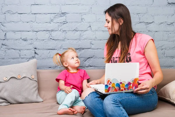 Молода мати і дочка двох років блондинка використовують ноутбук комп'ютерний ноутбук білий з яскравим принтом, сидячи на дивані в приміщенні — стокове фото