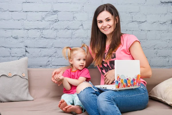 Mladá matka a dcera dvou let blondýnka použít přenosný počítač notebook bílé s jasně tisku sedí na gauči doma — Stock fotografie