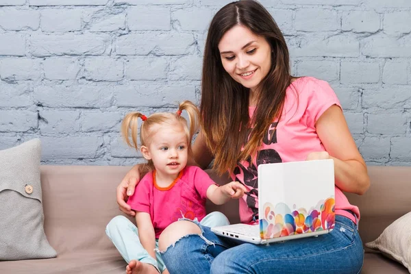 Молода мати і дочка двох років блондинка використовують ноутбук комп'ютерний ноутбук білий з яскравим принтом, сидячи на дивані в приміщенні — стокове фото