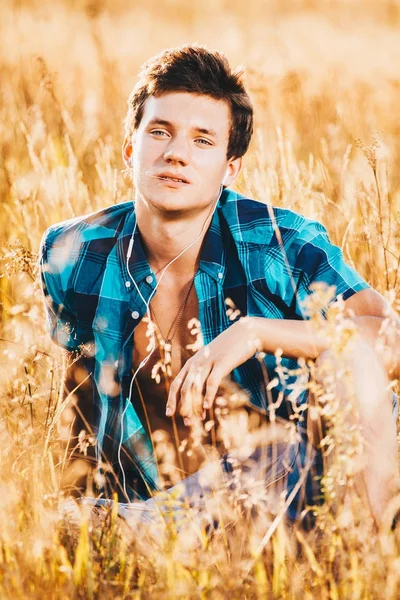 Beautiful sexy guy in an unbuttoned shirt and naked torso sits in a yellow field with spikelets and listens music in white headphones — Stock Photo, Image