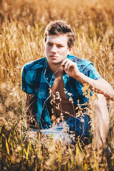 Bel ragazzo sexy in camicia sbottonata e torso nudo si siede in un campo giallo con spighette e ascolta musica in cuffie bianche — Foto Stock