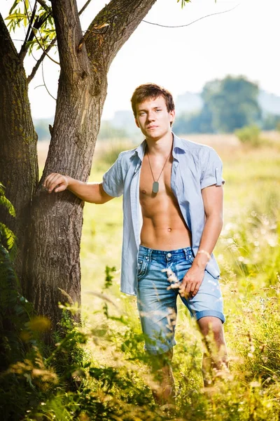 Νέων όμορφος άντρας σέξι με ένα γυμνό κορμό με ελαστικό κοιλιά μυών σε ένα ξεκούμπωτο πουκάμισο που ποζάρουν στη φύση σε ένα λιβάδι κοντά σε ένα δέντρο. — Φωτογραφία Αρχείου