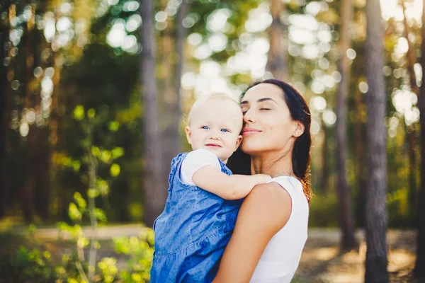 young beautiful fashionable mother with long brunette hair holding a daughter's blonde with blue eyes one year of birth in a coniferous forest in a summer park.