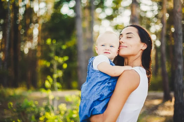 young beautiful fashionable mother with long brunette hair holding a daughter's blonde with blue eyes one year of birth in a coniferous forest in a summer park.