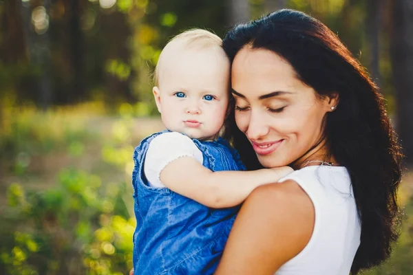 Junge schöne modische Mutter mit langen brünetten Haaren hält die Blondine einer Tochter mit blauen Augen ein Jahr nach ihrer Geburt in einem Nadelwald in einem Sommerpark. — Stockfoto