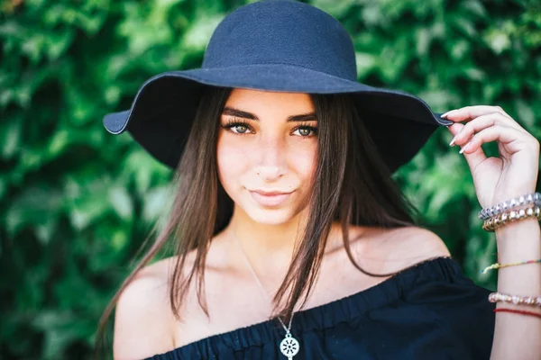 Beautiful young sexy girl with swarthy skin and brunette with black hair dressed in a stylish black dress and hat on background of green foliage — Stock Photo, Image