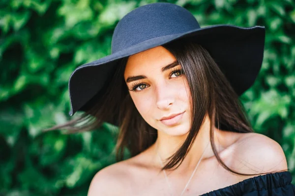 Bela jovem menina sexy com pele morena e morena com cabelo preto vestido com um vestido preto elegante e chapéu no fundo da folhagem verde — Fotografia de Stock