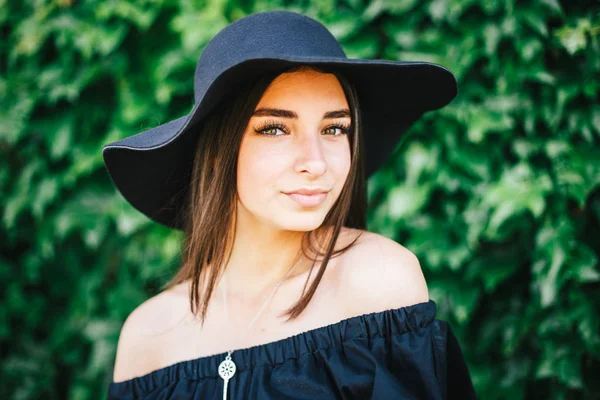 Beautiful young sexy girl with swarthy skin and brunette with black hair dressed in a stylish black dress and hat on background of green foliage — Stock Photo, Image