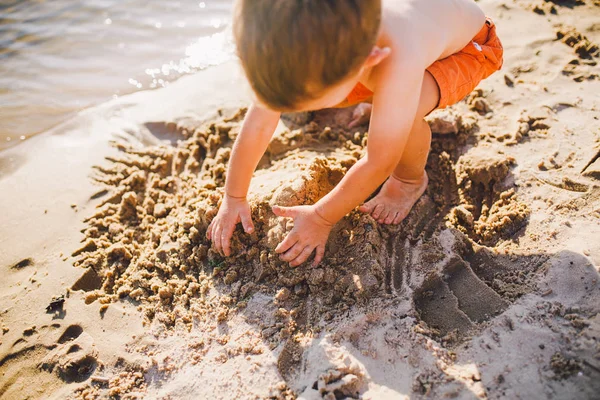 Un petit garçon construit des figures du sable sur le rivage de l'étang au coucher du soleil de la journée, les mains déterrer le sable dans un plan croustillant — Photo