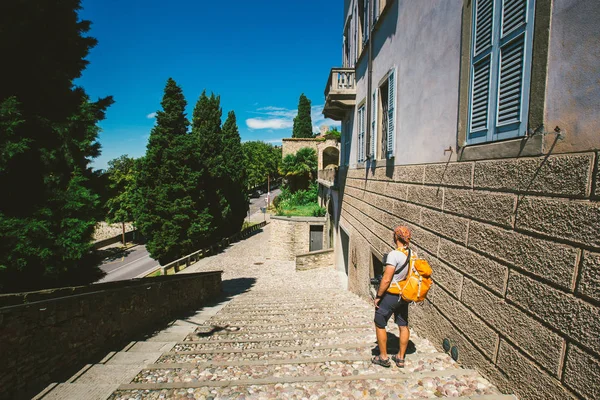 Homem do turista fica de costas para a antiga cidade europeia de Bérgamo, na Itália, no verão. Vestida com um xale e uma camiseta com uma mochila laranja e um lenço na cabeça em suas mãos segura — Fotografia de Stock