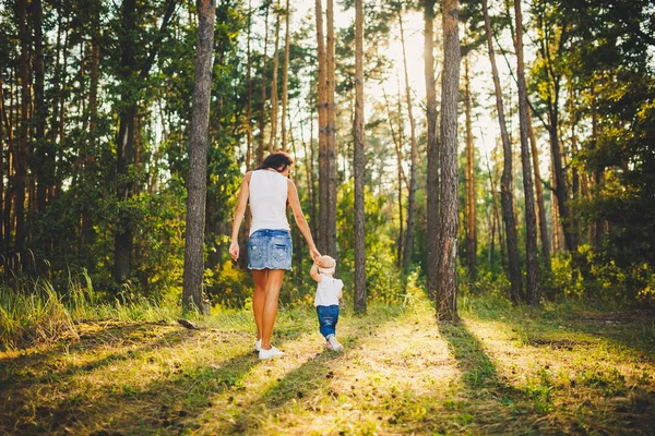 beautiful and stylish mother leads the hand of a girl of one year from birth in the park against the background of tall coniferous trees. Family values mom learns to walk child