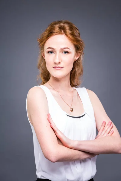 Retrato de una joven pelirroja hermosa chica en el estudio sobre un fondo gris aislado. Una mujer está de pie con los brazos cruzados y sonriendo con una camisa blanca con una manga corta. Concepto empresarial — Foto de Stock