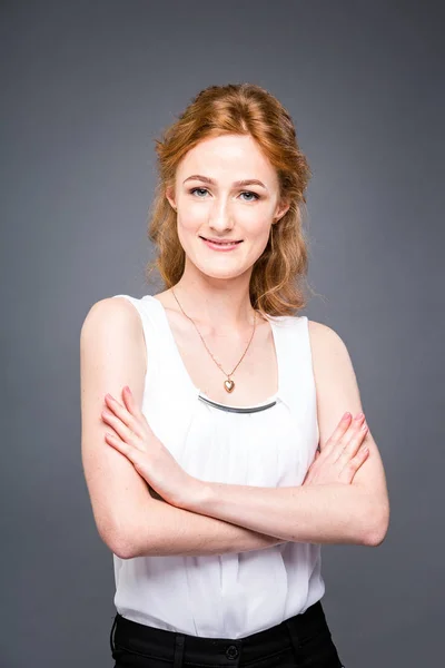 Retrato de una joven pelirroja hermosa chica en el estudio sobre un fondo gris aislado. Una mujer está de pie con los brazos cruzados y sonriendo con una camisa blanca con una manga corta. Concepto empresarial — Foto de Stock