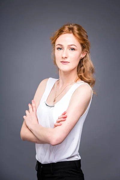 Retrato de una joven pelirroja hermosa chica en el estudio sobre un fondo gris aislado. Una mujer está de pie con los brazos cruzados y sonriendo con una camisa blanca con una manga corta. Concepto empresarial — Foto de Stock